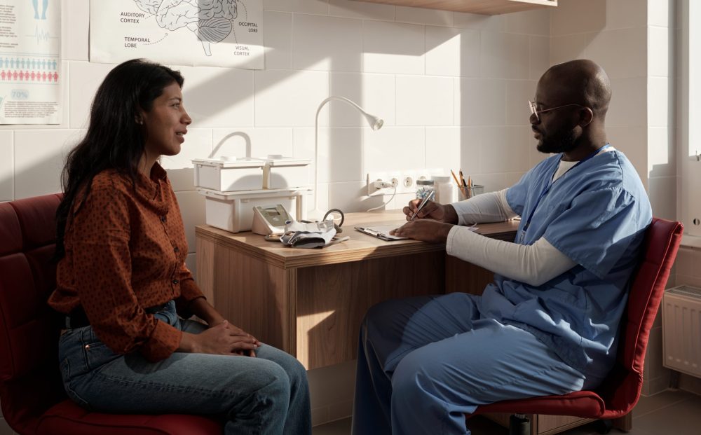 Young female patient sitting in front of doctor making prescription notes and describing him symptoms of her ailment in hospital
