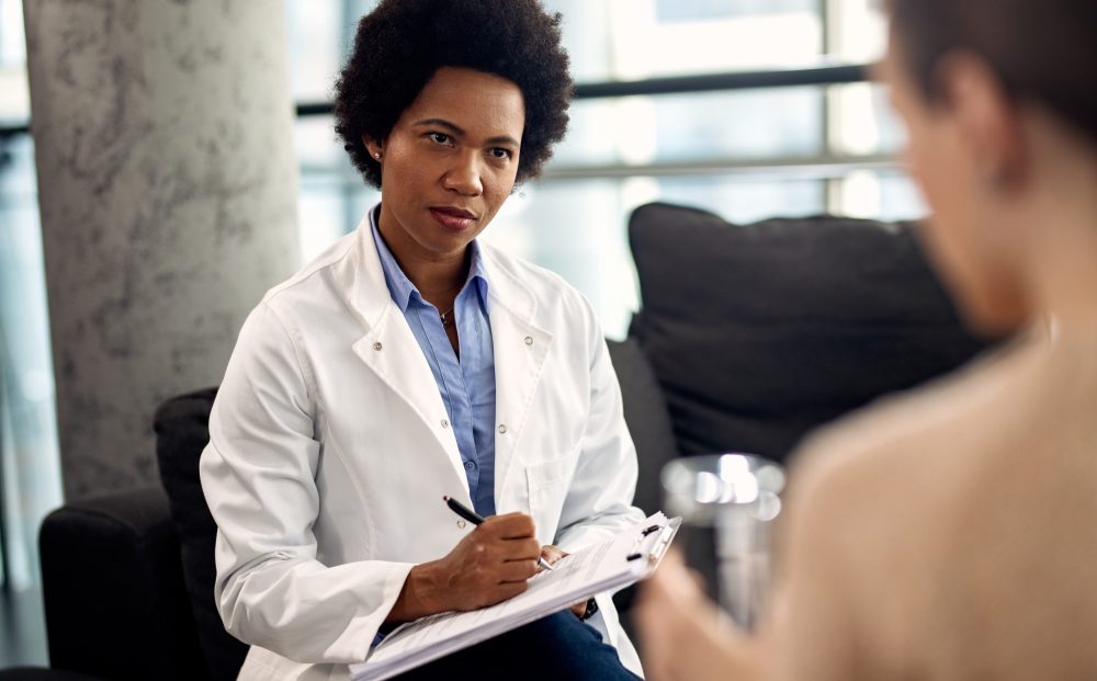 African American doctor writing notes while listening to patient during a psychotherapy session.