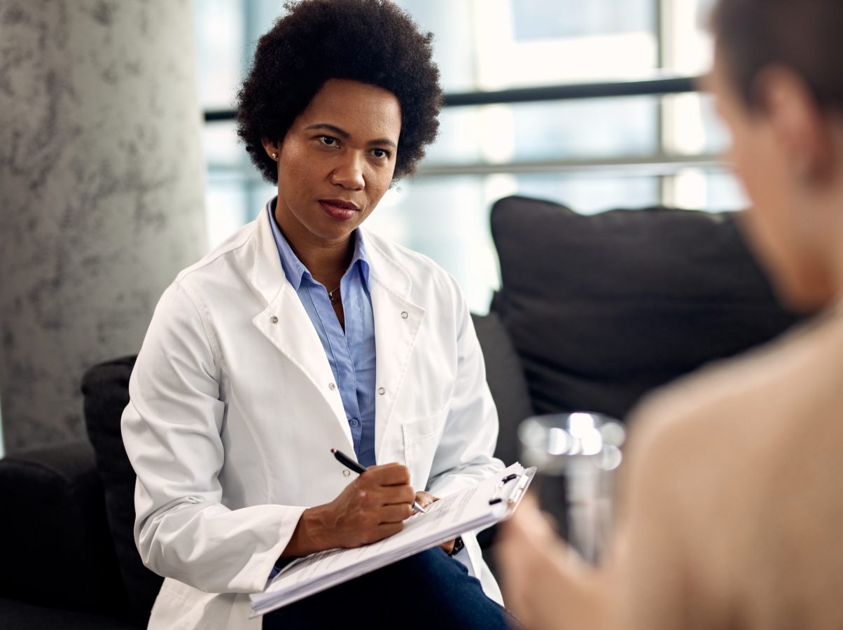 African American doctor writing notes while listening to patient during a psychotherapy session.