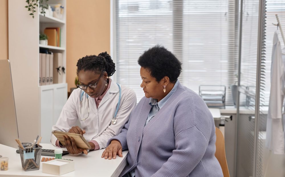 Medium shot of African American female doctor and senior patient looking at tablet computer with medical results at table in clinic