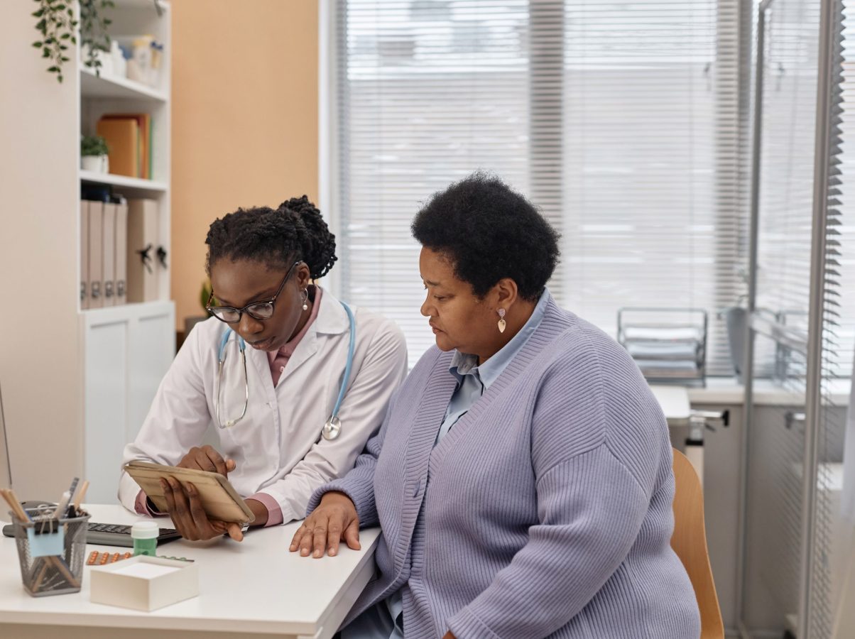 Medium shot of African American female doctor and senior patient looking at tablet computer with medical results at table in clinic