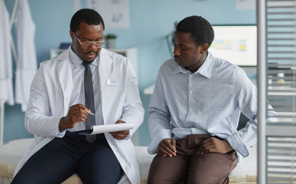 Black middle aged male doctor explaining medical information to young Black man patient during consultation in clinic office, both sitting on examination table and discussing health