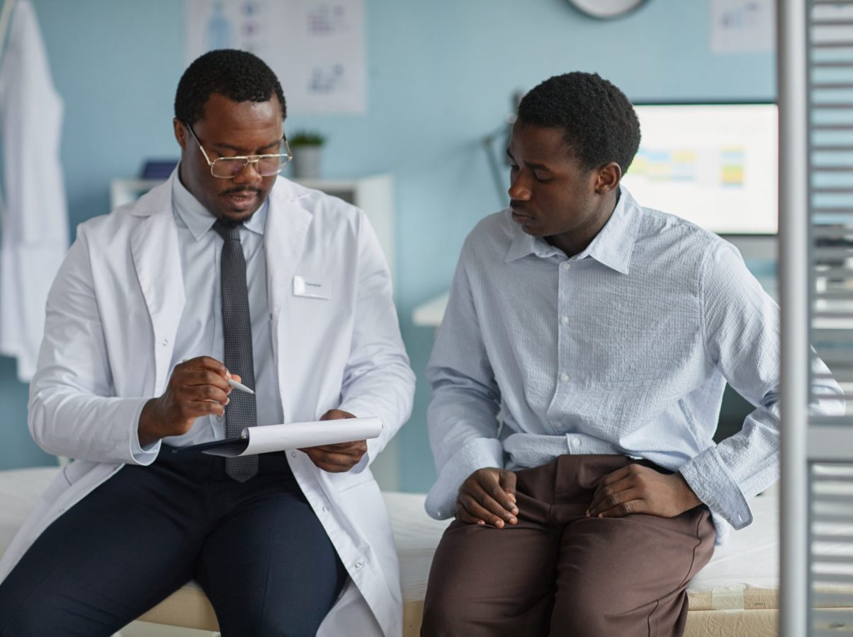 Black middle aged male doctor explaining medical information to young Black man patient during consultation in clinic office, both sitting on examination table and discussing health