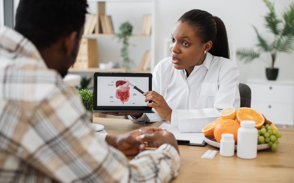 African american specialist in nutrition holding tablet with gastrointestinal tract on screen while counseling man at work. Focused woman explaining client about food intolerance during appointment.