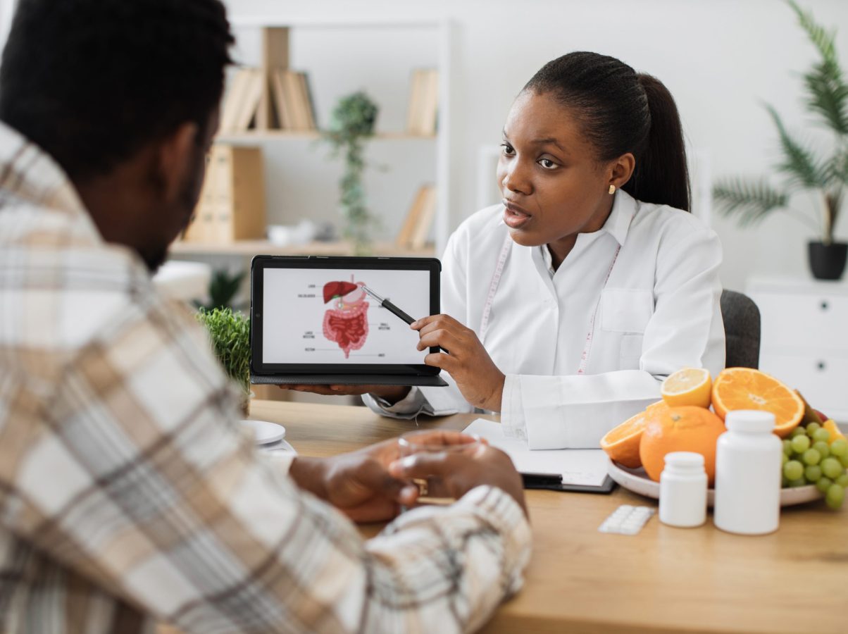 African american specialist in nutrition holding tablet with gastrointestinal tract on screen while counseling man at work. Focused woman explaining client about food intolerance during appointment.