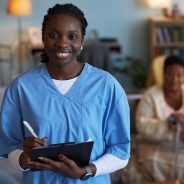 Portrait of young adult Black woman wearing medical scrubs smiling at camera holding clipboard and pen, senior Black woman sitting in background using walking cane in living room, during patient visit