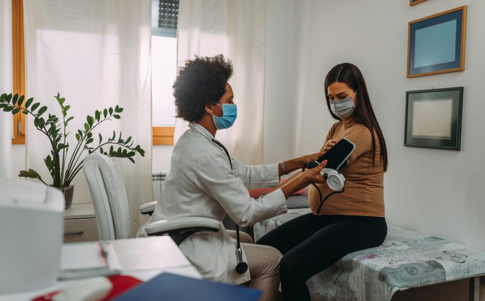 A pregnant woman during a routine check up with her doctor. Doctor is measuring pressure to her while they are wearing face masks