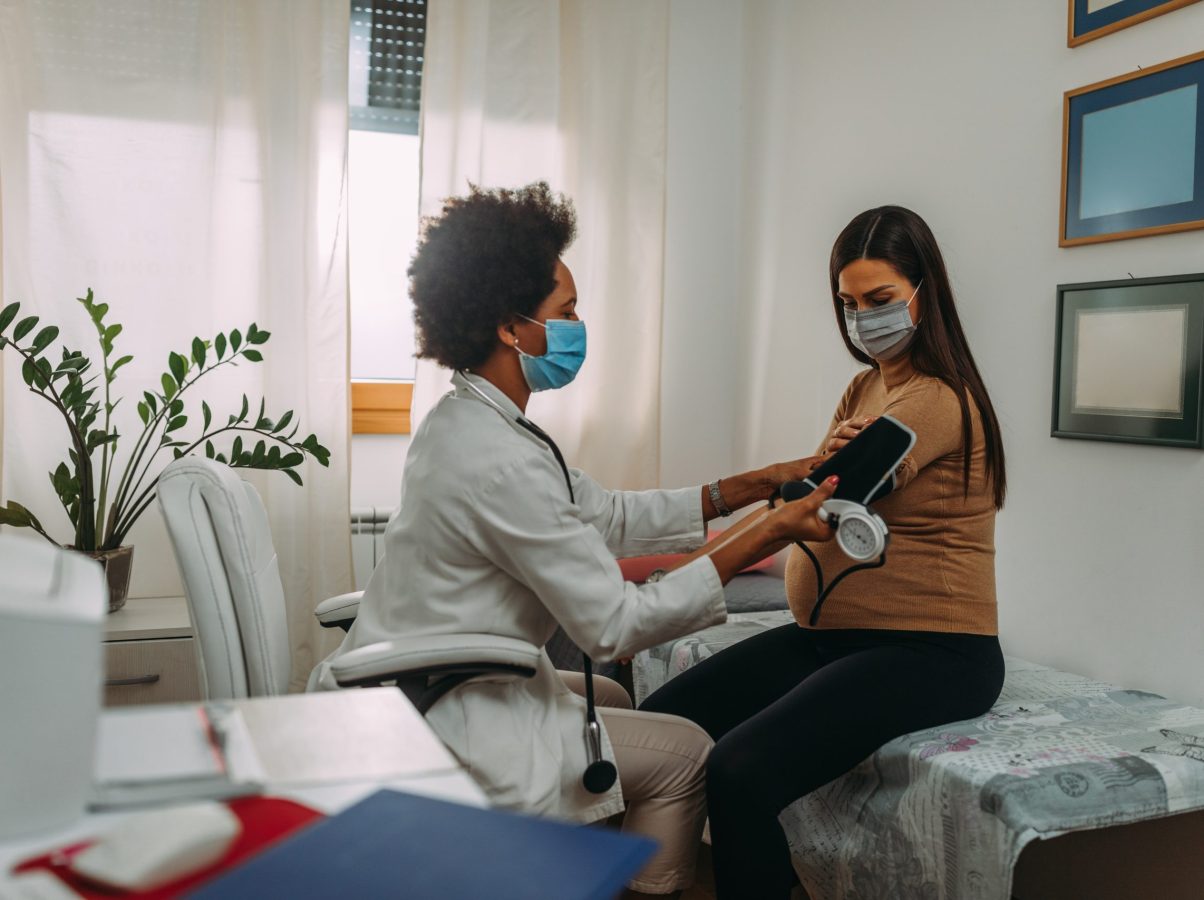 A pregnant woman during a routine check up with her doctor. Doctor is measuring pressure to her while they are wearing face masks